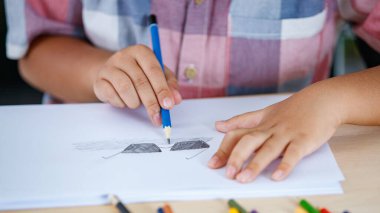 Pembe tişörtlü küçük Asyalı çocuk sanat dersi almak için renkli kalemlerle dolu sınıfta oturuyor ve beyaz kağıda mavi kalemle siyah gözlük resmi çiziyor ve gölgeliyor.