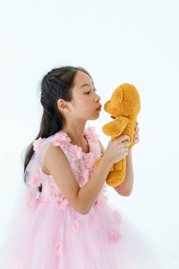 Asyalı güzel kızın portre resmi beyaz prenses çiçekli elbise giyer. Moda kostümü. Kameraya bak ve beyaz arka planda pofuduk turuncu oyuncak ayıya sarıl..