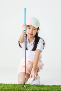 Portre, Asyalı küçük profesyonel golfçünün izole edilmiş, spor kıyafetli küçük kız golfçüsünün yeşil çimenlere diz çöktüğü ve golf sopası tutarken beyaz arka planda topu gösterdiği yakın çekim..