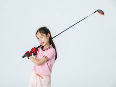 Stüdyoda, uzun saçlı, spor üniformalı ve renkli şapkalı küçük Asyalı golfçünün beyaz arka planda golf sürücüsü pozu..