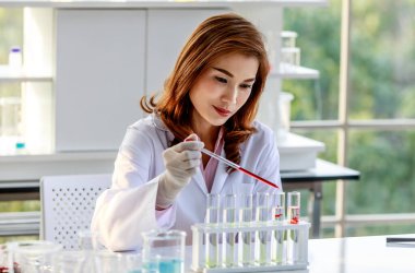 Laboratuvarda araştırma yaparken laboratuvar önlüğü ve eldivenleriyle renkli kimyasal sıvıları şişelerde karıştıran odaklanmış genç Asyalı kadın kimyacı..