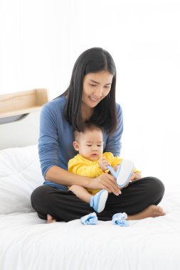 Asyalı kadın için portre dikey fotoğraf anne gülümsüyor ve beyaz yatakta bacağında da bebek oturuyor. Bebek sabahları yatak odasında çorapla oynuyor..