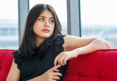 Genç Taylandlı bir gencin kırmızı koltukta oturup pencereden dışarı bakarken çekilmiş yakın plan portresi. Genç kız gözleri üzgün ve yalnız görünüyor.