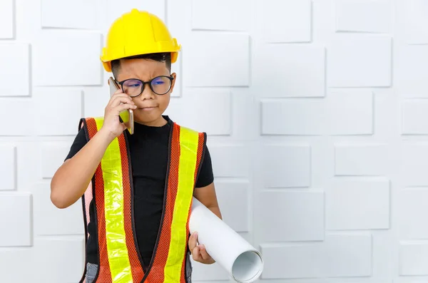 作为一名工程师 年轻的亚洲男孩戴着眼镜 黑色衬衫 黄色的安全背心和头盔 拿着纸卷 站在白墙边 一边用智能手机打电话寻找有疑问的工作 — 图库照片