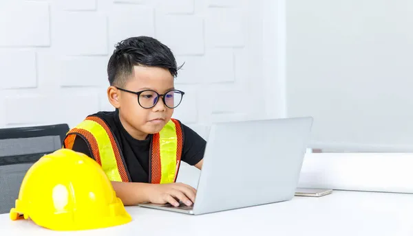 聪明的年轻亚洲男孩 戴着眼镜和黄色的安全背心 坐在白色的写字台上 紧紧抓住下巴 在保护头盔旁边认真地工作 以考虑困难的工作 — 图库照片