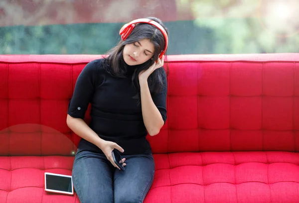 赤いソファに座ってヘッドセットで音楽を聴いている若いタイ トルコの10代の肖像写真 手で音楽を楽しむカジュアルな服を着たジュニア女の子がヘッドフォンジェスチャーに触れる — ストック写真