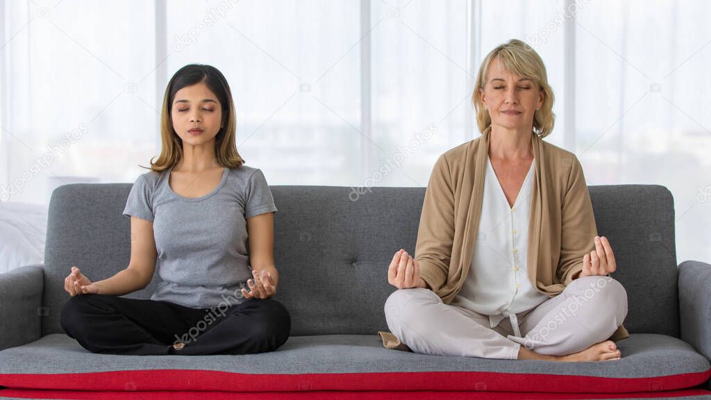 Full body of middle aged mother in law and young Asian daughter in law sitting on sofa in lotus pose and meditating together at home