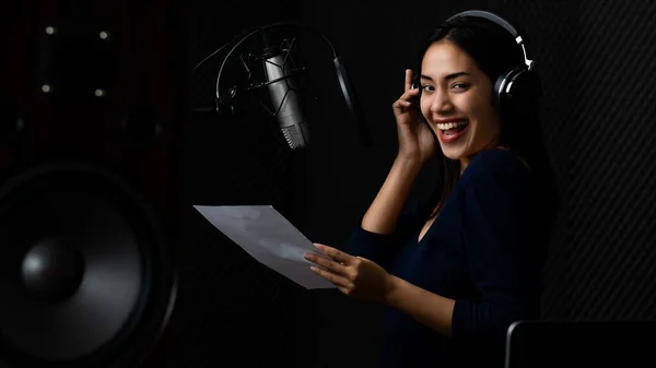 Beautiful Asian woman singer holding lyrics paper and wears headphone with smile while enjoy practicing song entertainment by recording elegant voice to microphone in dark audio room of music studio.