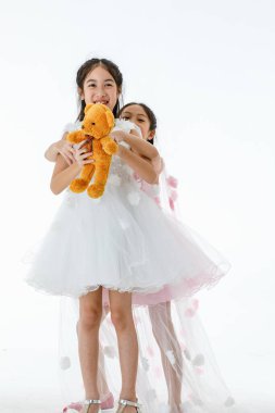 Portre, iki küçük Asyalı balerin, pembe ve beyaz güller içinde bale elbisesi giyip turuncu oyuncak ayı bebeği için gülerken çekilmiş..
