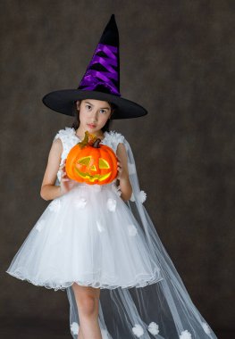 Küçük Asyalı şirin kızın, uzun şapka giymiş, gülümseyen ve elinde turuncu balkabağıyla Cadılar Bayramı Festivali 'nde şeker mi şaka mı oynamaya hazır bir şekilde çekilmiş portresi..