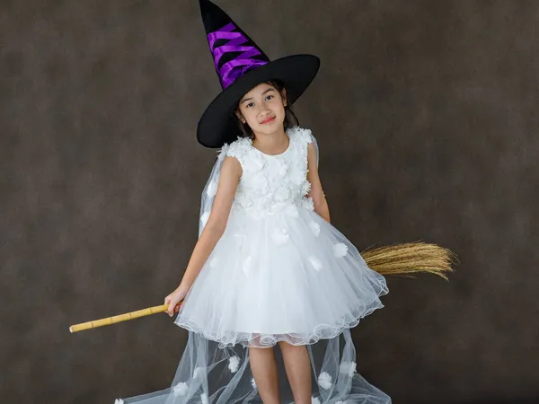 白い魔女の衣装で小さなかわいいアジアの子供のポートレートスタジオショット黒紫色の高い帽子立っている彫刻オレンジカボチャの顔と暗い背景の前に魔法のブロムスティック — ストック写真