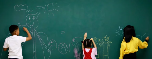 アジアの小さな幸せなスマート小学生と女子学生は テーブルと椅子でいっぱいの教室の前で緑の黒板にチョーク書きの漫画のキャラクターを書くことを楽しんでいます — ストック写真