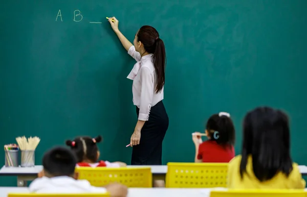 アジアの若い美しい女性の肖像画英語教授の講師は 小学校の教室で生徒のための黒板にアルファベットの質問を書くチョークを使用して立っている — ストック写真