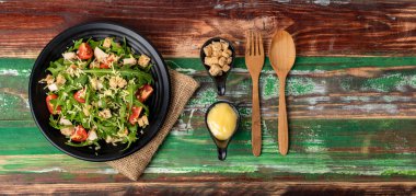 Domatesli roket salatası, kızarmış tavuk, çuval bezinde parmesan peyniri. Tahta çatal bıçak ve mayonez ve fazladan ekmek dolu iki küçük sos kabı ağaçlık masada..