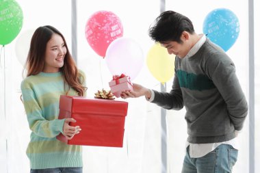 Güzel genç Asyalı kadın mutlu bir şekilde gülümsüyor ve kışın coşkulu aşkı kutlamak için süslü balonlarla süslenmiş romantik kapalı hava partisinde sevgili erkek arkadaşından güzel bir kırmızı hediye kutusu aldığına seviniyor.