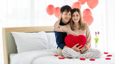 İyimser genç Asyalı çift, kalp şeklinde yastıkları var. Gülümsüyorlar ve kameraya bakıyorlar. Yatakta birbirlerine sarılıyorlar.