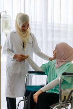 Genç Müslüman kadın doktor konuşuyor ve hastanede tekerlekli sandalyede oturan hasta bir müslümanı neşelendiriyor..