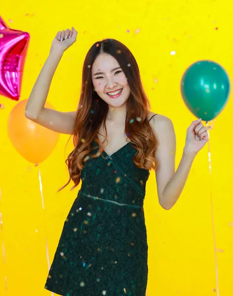 stock image Asian happy girl wears sexy dress standing posing and fun excited with shiny paper confetti in hand in celebration party in front yellow wall background and colorful helium air balloons.