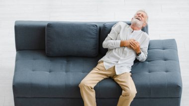 Beyaz, yaşlı, yaşlı, yaşlı, kır saçlı, sakallı bir adam. Oturma odasındaki koltuğa uzanmış, tek başına göğsünde el ele tutuşmuş. Acil kalp krizi geçiriyor..