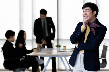 LGBTQ transseksüel ofis çalışanının portresi sıradan takım elbiseli ve kamera önünde neşeli ve mutlu bir hareketle arka planda iş arkadaşları bulanıklaşır.