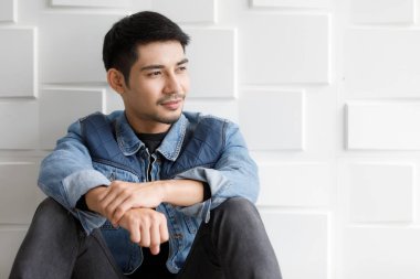 Cilveli kot ceketli Asyalı genç adam beyaz duvara karşı dost canlısı ve gülümseyen hareketlerle oturuyor. Moda şehir portresi konsepti
