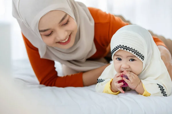 一位漂亮可爱的亚洲穆斯林 身穿头巾裙 与女儿玩耍 与床上的奶嘴玩耍 — 图库照片