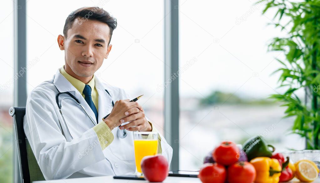 Asian male nutritionist clasping hands and looking at camera while sitting at desk with healthy food in office of modern hospital