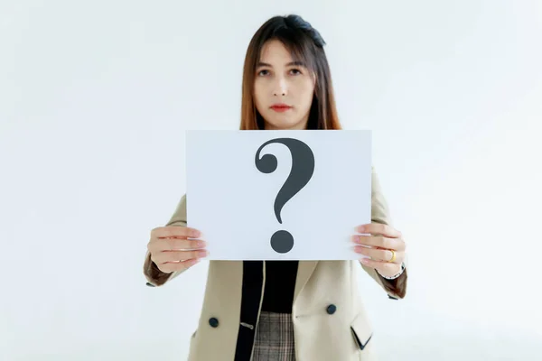 Studio shot of doubtful female officer staff in business suit look at camera holding question mark paper cardboard sign showing curiosity thinking for answers on white background.