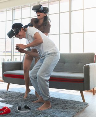 Genç ve çekici Asyalı çift ellerinde joysticklerle birlikte hoplayıp zıplamaktan ve video oyunları oynamaktan zevk alıyorlar. Oturma odasında cam pencereli eğlence ve heyecan dolu gözlükler takıyorlar..