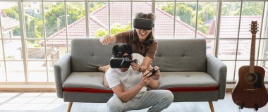 Genç ve çekici Asyalı çift ellerinde joysticklerle birlikte hoplayıp zıplamaktan ve video oyunları oynamaktan zevk alıyorlar. Oturma odasında cam pencereli eğlence ve heyecan dolu gözlükler takıyorlar..