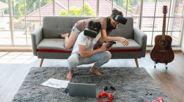 Genç ve çekici Asyalı çift ellerinde joysticklerle birlikte hoplayıp zıplamaktan ve video oyunları oynamaktan zevk alıyorlar. Oturma odasında cam pencereli eğlence ve heyecan dolu gözlükler takıyorlar..