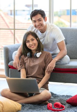 Genç ve çekici Asyalı çift oturma odasında oturup dizüstü bilgisayara bakıyorlar. Kadın yerde kulaklık takmış, adam kanepede yatıyor. Aşk ve çift ilişkisi kavramı.