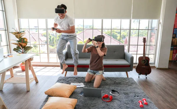 Genç ve çekici Asyalı çift ellerinde joysticklerle birlikte hoplayıp zıplamaktan ve video oyunları oynamaktan zevk alıyorlar. Oturma odasında cam pencereli eğlence ve heyecan dolu gözlükler takıyorlar..