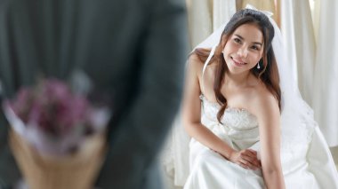 Asyalı, güzel, mutlu, uzun saçlı, beyaz gelinlikli, dantelli duvağı olan bir gelin. Gri takım elbiseli bir damat bekliyor. Çiçek buketini arkasına saklayıp sürpriz yapmak için soyunma odasında bekliyor..