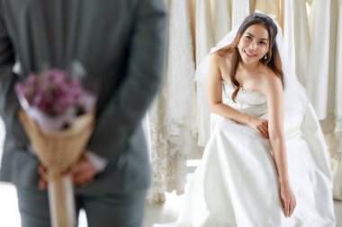 Asian young beautiful happy long hair bride in white wedding dress with seethrough lace veil sit smile waiting for groom in gray suit who hide flower bouquet behind back to surprise in dressing room. clipart