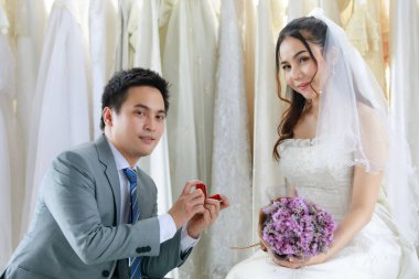 Gri takım elbiseli Asyalı yakışıklı damat kırmızı elmas yüzük kutusunu tutuyor. Beyaz gelinlik içinde, elinde çiçek buketi tutan güzel bir gelini, soyunma odasında sunuyor..