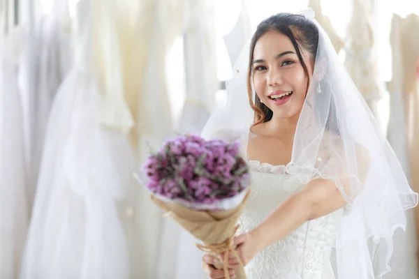 白いドレスのアジアの美しい花嫁とともにシースルーヘアレースベール立って笑みを浮かべて見てカメラを保持紫色の香りの結婚式の花花束で手の中にぼやけたフォアグラウンドで脱衣所 — ストック写真