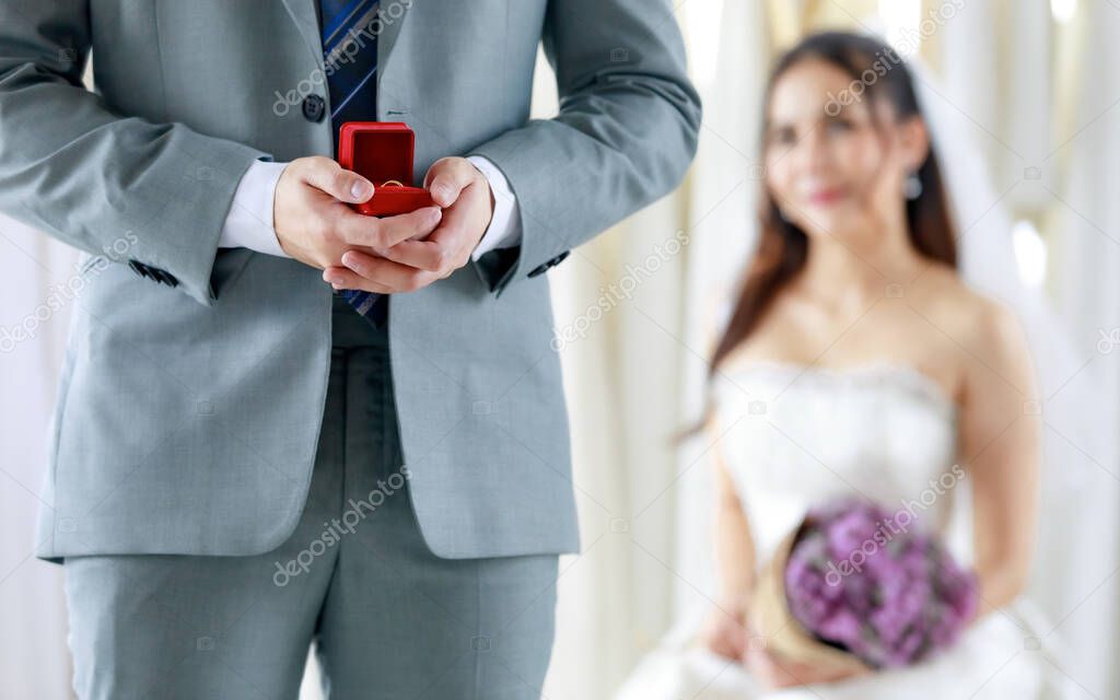 Unrecognizable groom in grey formal suit stand hold red box of diamond ring prepare giving to Asian young beautiful happy bride in white wedding dress holding flower bouquet in blurred background.
