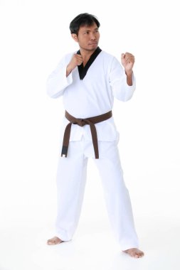 Asyalı orta yaşlı erkek tekvando karate sporcusu üniformalı, kahverengi kemerli beyaz arka plan önünde güçlü bir hareket sergileyen kameraya bakarken çekilmiş tek bir portre resmi..
