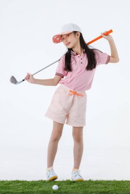 Stüdyoda, uzun saçlı, spor üniformalı ve renkli şapkalı küçük Asyalı golfçünün beyaz arka planda golf sürücüsü pozu..
