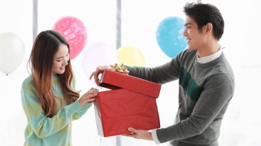 Güzel, Asyalı bir kadın sürpriz bir hediyeyle heyecanlanıyor. Kırmızı kutunun içinde sevgili nişanlısının kış mevsiminde coşkulu bir aşkı kutlamak için süslü balonlarla süslenmiş kapalı mekan partisinde açtığı sürpriz hediye.