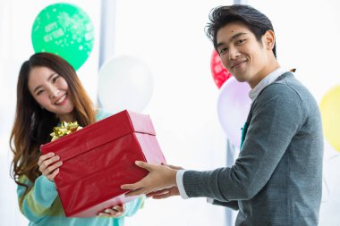 İki Asyalı aşık, kışın coşkulu aşklarını kutlamak için süslü balonlarla süslenmiş kapalı mekan partisinde mutlu yüzlerle hediye alışverişi yapıyorlar.