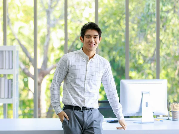 Asyalı başarılı beyaz yakalı erkek memurun, çalışma masasına yaslanıp bilgisayar monitörüyle gülümseyen kameraya bakıp elini pantolonunun cebine soktuğu bir portre resmi..