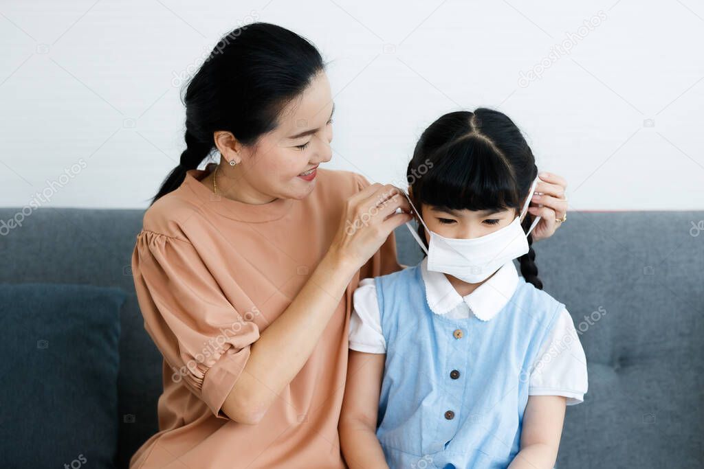 Close up shot of Asian lovely mother sitting on sofa in living room helping little cute daughter in school uniform wearing safety hygiene protecting face mask preventing coronavirus during outbreak.