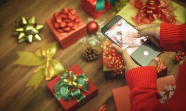 Noel partisinin büyüleyici dekorasyonunu ezberlemek için güzel renkli fiyonklu ve süslemeli lüks hediye kutusunun fotoğrafını çekmek ve kaydetmek için akıllı telefon kullanan kişi.