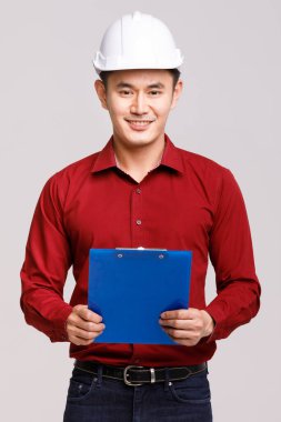 Asyalı olumlu mühendis kasketli ve gri arka planda duran panosuyla stüdyodaki kamerayı işaret ediyor.