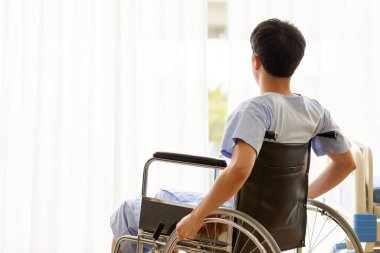 Mavi hastane üniformalı Asya 'lı, umut dolu bir erkek hasta, tekerlekli sandalyede oturur, sıcak ışıkta koğuş odasının beyaz perdesinden dışarı bakar ve ciddi yaralanma bacak probleminden kurtulmayı umut eder..