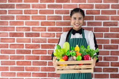 Yeşil önlüklü Asyalı mutlu kadın çiftçi gülümse. Kameraya bak. Elinde çiğ organik sebzelerle dolu tahta sepet var..