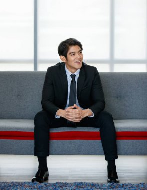 Yakın plan 20 'li yaşlarda Asyalı genç ve çekici bir iş adamı resmi bir kravat takıyor, rahat bir koltukta oturuyor ve ofisin içinde rahatlıyor, güven ve mutlulukla gülümsüyor.