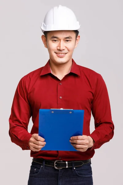 正面的亚洲男性工程师承包商 戴着硬礼帽 剪贴板站在灰色背景上 指着工作室里的相机 — 图库照片